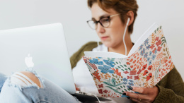 Eine Frau sitzt am Laptop, in ihrer Hand ein Heft mit buntem Umschlag.