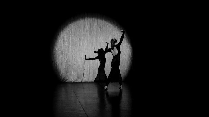 Ballettänzerin in einem schwarzen Abendkleid auf einer Bühne, von einem einzelnen Scheinwerfer beleuchtet (schwarzweiß)
